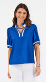 A woman modeling a royal blue short-sleeve blouse with white trim on the collar and cuffs, paired with white pants, against a plain white studio background.