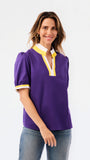 A woman modeling a purple short-sleeve blouse with a white and yellow V-neck collar and yellow striped cuffs, paired with blue jeans, against a plain white background.