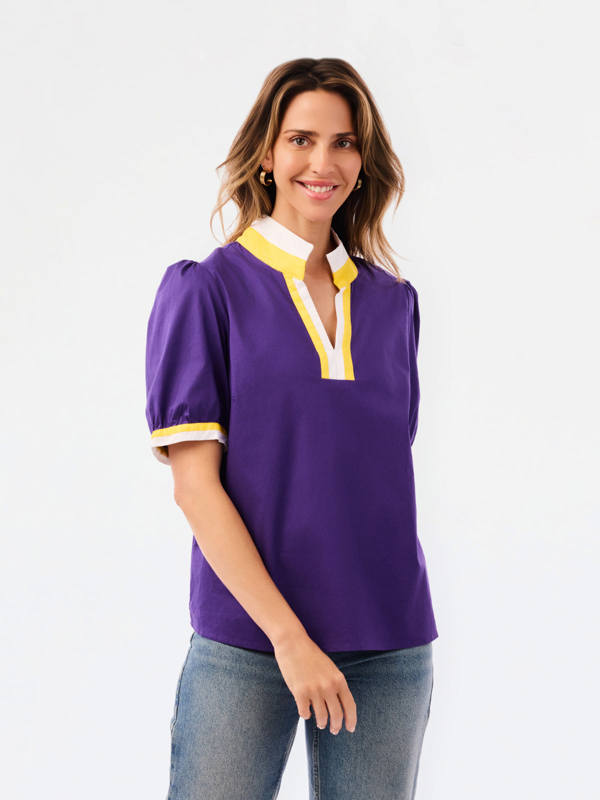 A woman modeling a purple short-sleeve blouse with a white and yellow V-neck collar and yellow striped cuffs, paired with blue jeans, against a plain white background.