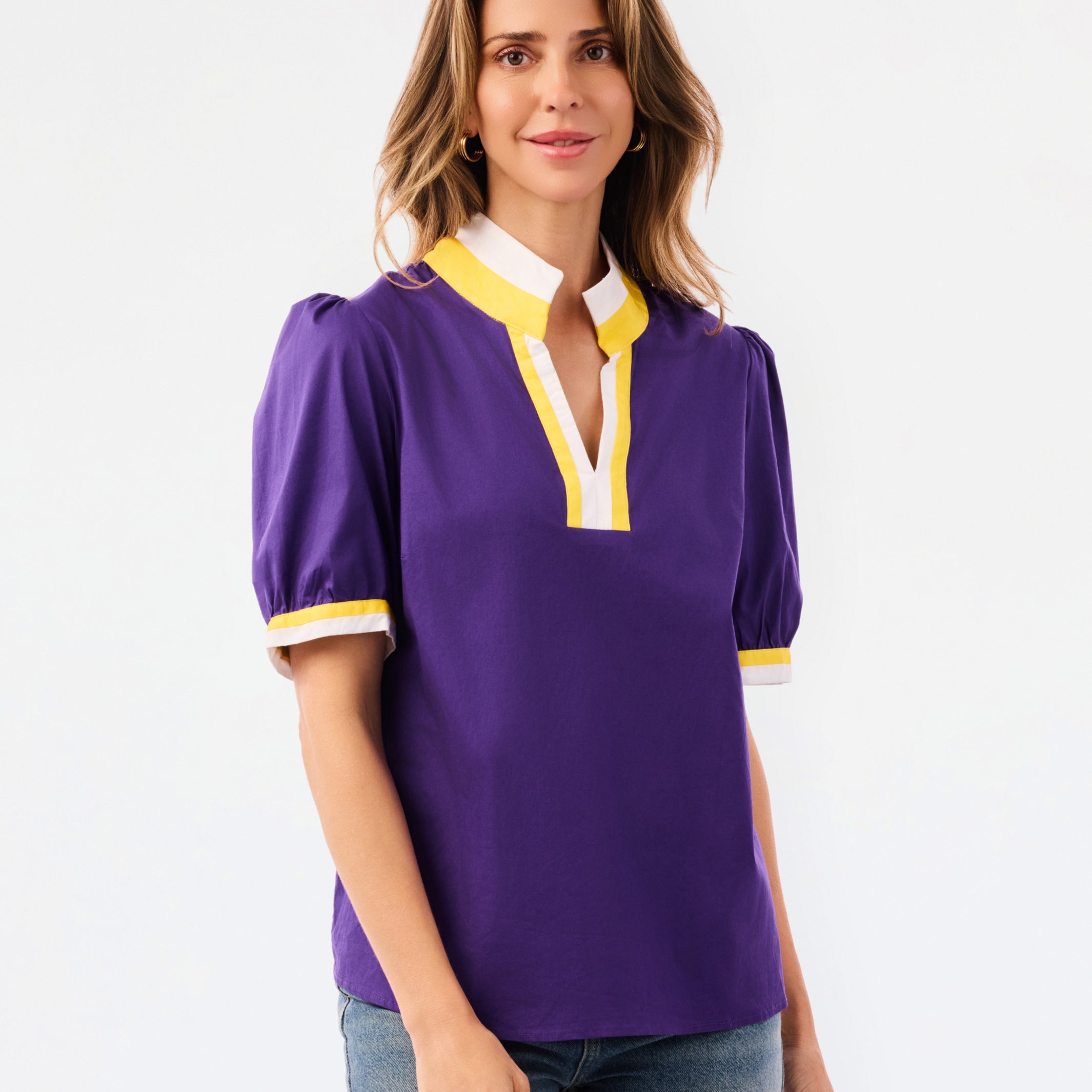 A woman modeling a purple short-sleeve blouse with a white and yellow V-neck collar and yellow striped cuffs, paired with blue jeans, against a plain white background.