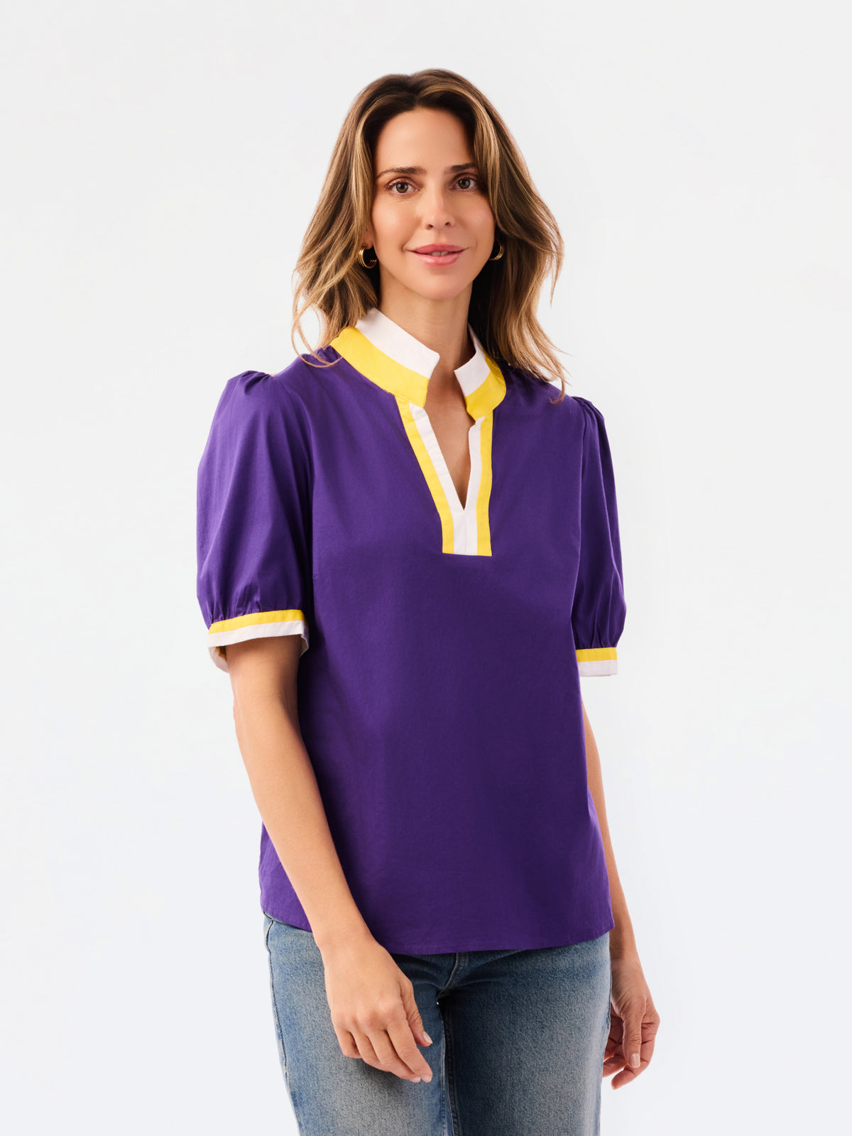 A woman modeling a purple short-sleeve blouse with a white and yellow V-neck collar and yellow striped cuffs, paired with blue jeans, against a plain white background.