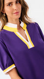 A woman modeling a purple short-sleeve blouse with a white and yellow V-neck collar and yellow striped cuffs, paired with blue jeans, against a plain white background.