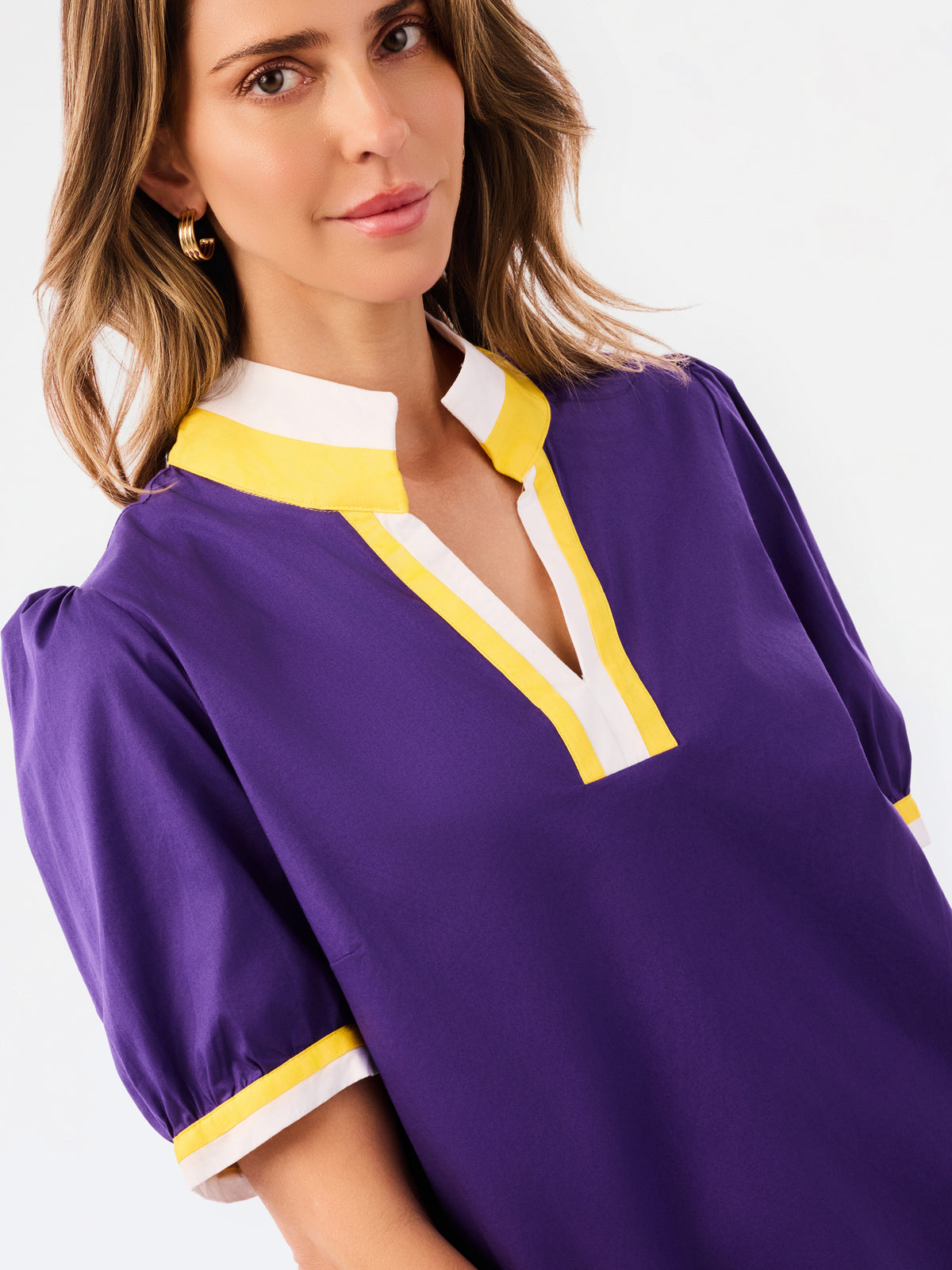 A woman modeling a purple short-sleeve blouse with a white and yellow V-neck collar and yellow striped cuffs, paired with blue jeans, against a plain white background.