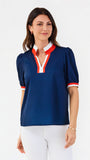 A woman modeling a navy blue short-sleeve blouse with red and white trim on the collar, placket, and sleeve cuffs, paired with white pants against a plain white studio background.