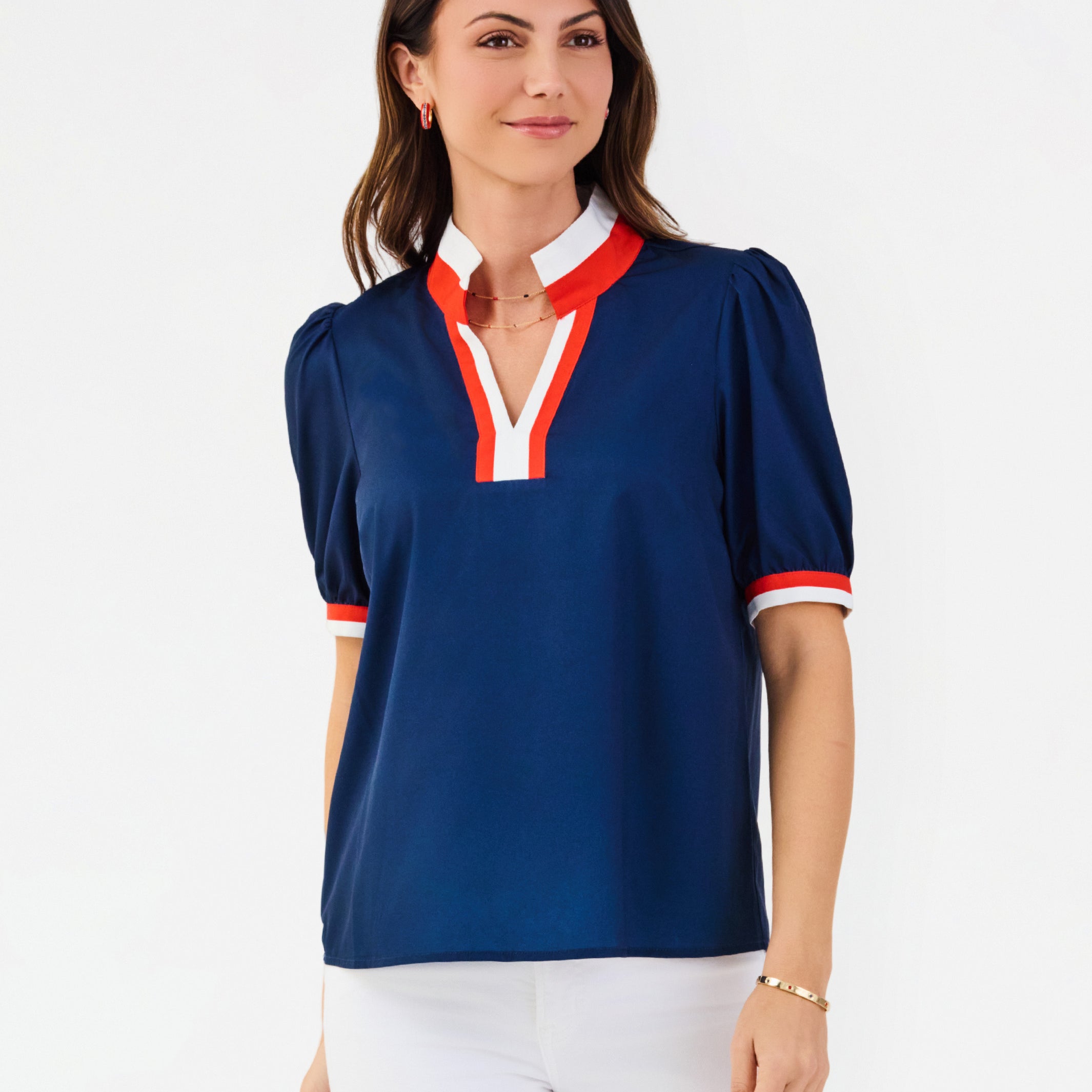 A woman modeling a navy blue short-sleeve blouse with red and white trim on the collar, placket, and sleeve cuffs, paired with white pants against a plain white studio background.