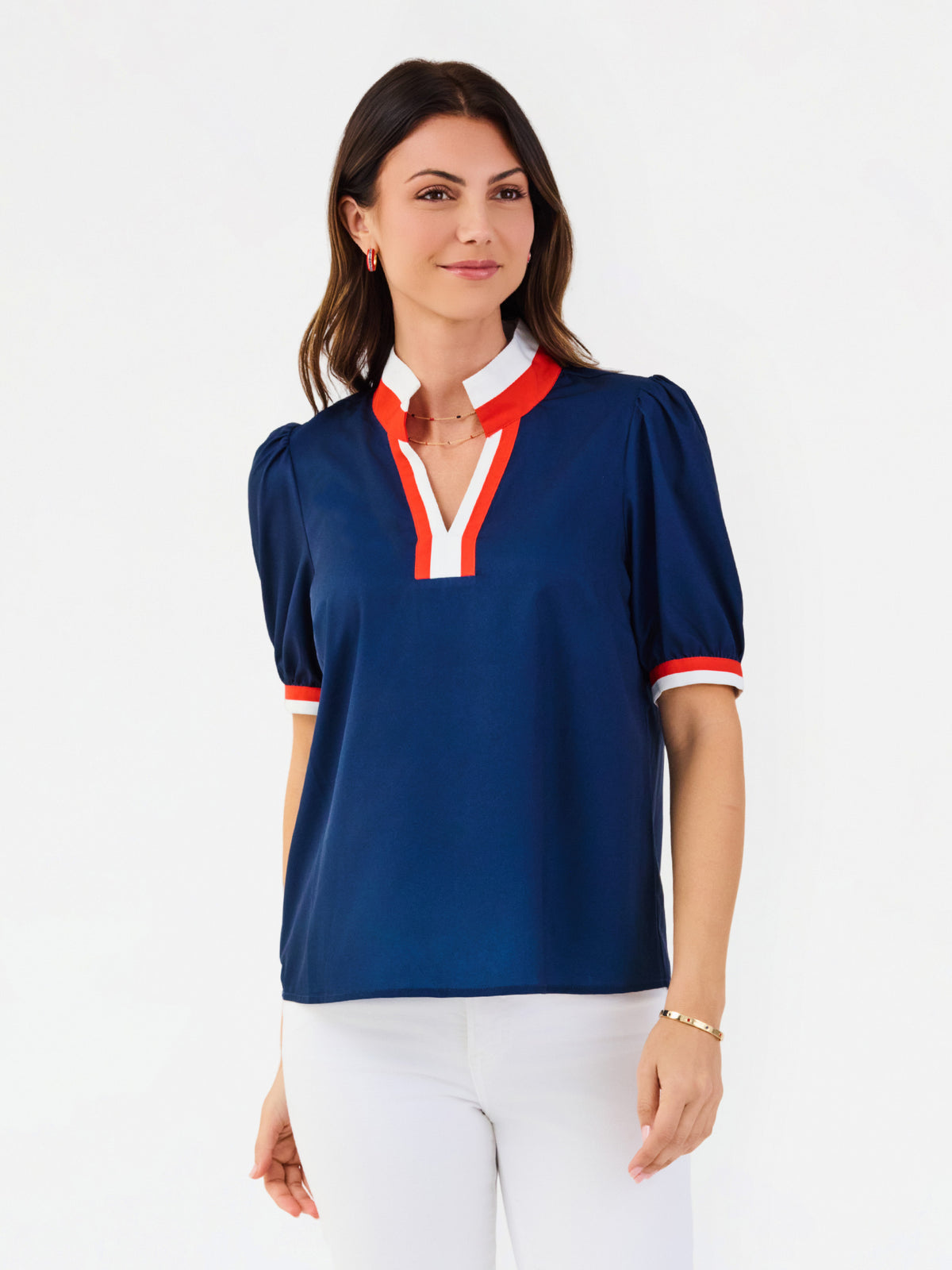 A woman modeling a navy blue short-sleeve blouse with red and white trim on the collar, placket, and sleeve cuffs, paired with white pants against a plain white studio background.