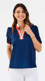 A woman modeling a navy blue short-sleeve polo shirt with a red and white V-neck trim, paired with white pants, against a plain white background.