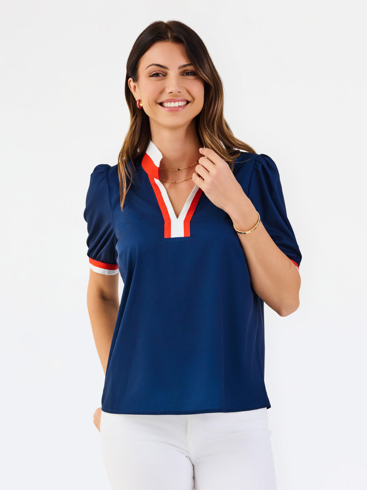 A woman modeling a navy blue short-sleeve polo shirt with a red and white V-neck trim, paired with white pants, against a plain white background.