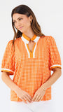 A woman modeling a short-sleeve orange and white gingham top with a white collar and orange trim, paired with white pants, against a plain white background