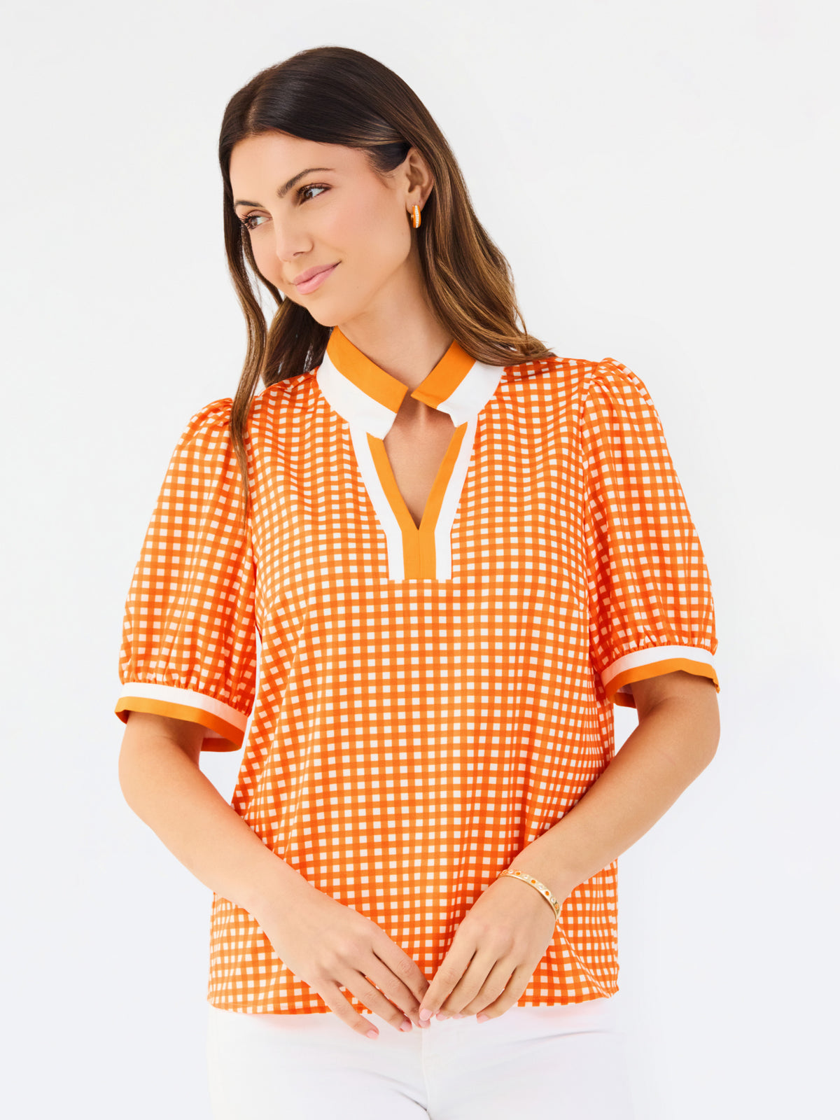 A woman modeling a short-sleeve orange and white gingham top with a white collar and orange trim, paired with white pants, against a plain white background