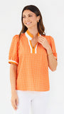 A woman modeling a short-sleeve orange and white gingham top with a white collar and orange trim, paired with white pants, against a plain white background.