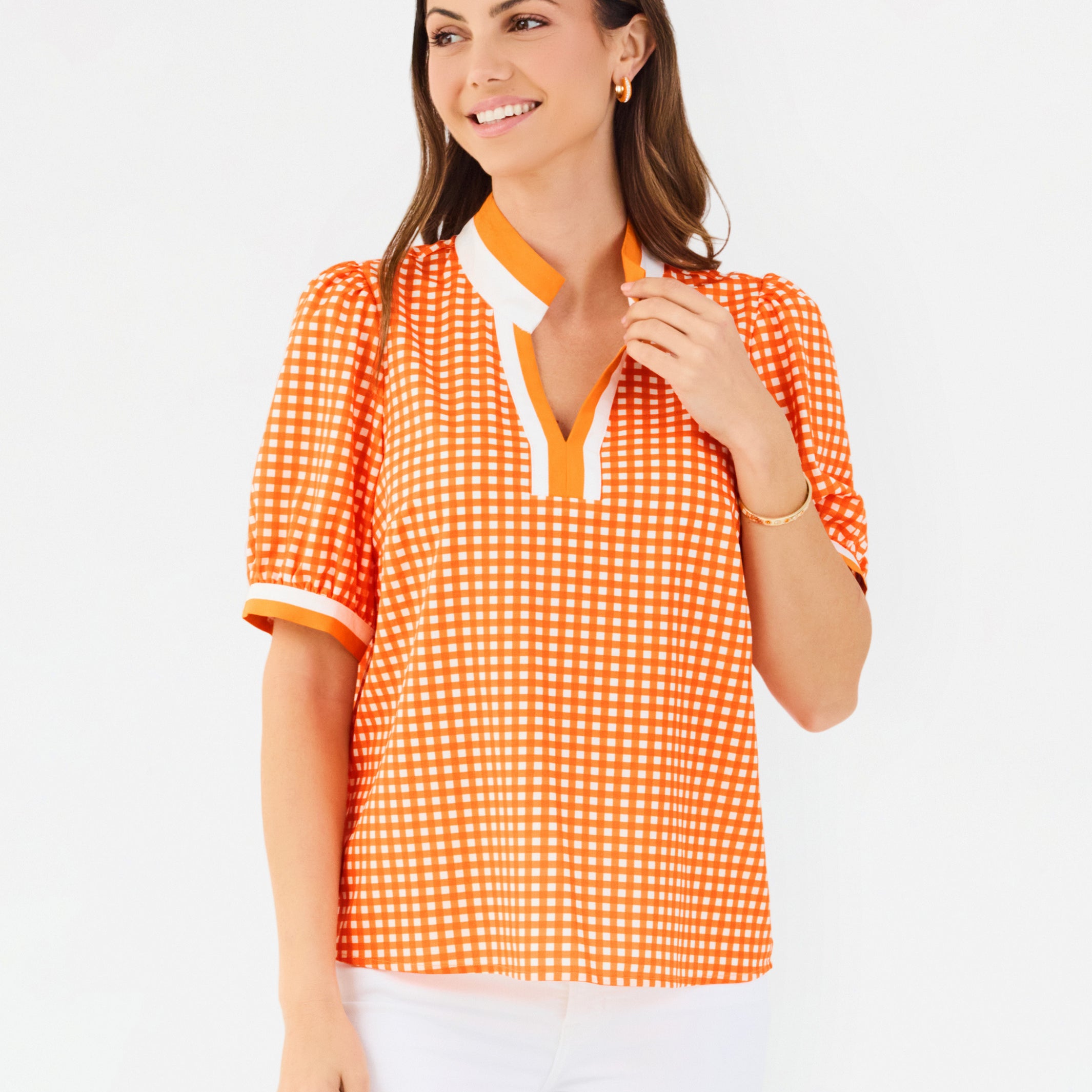 A woman modeling a short-sleeve orange and white gingham top with a white collar and orange trim, paired with white pants, against a plain white background.