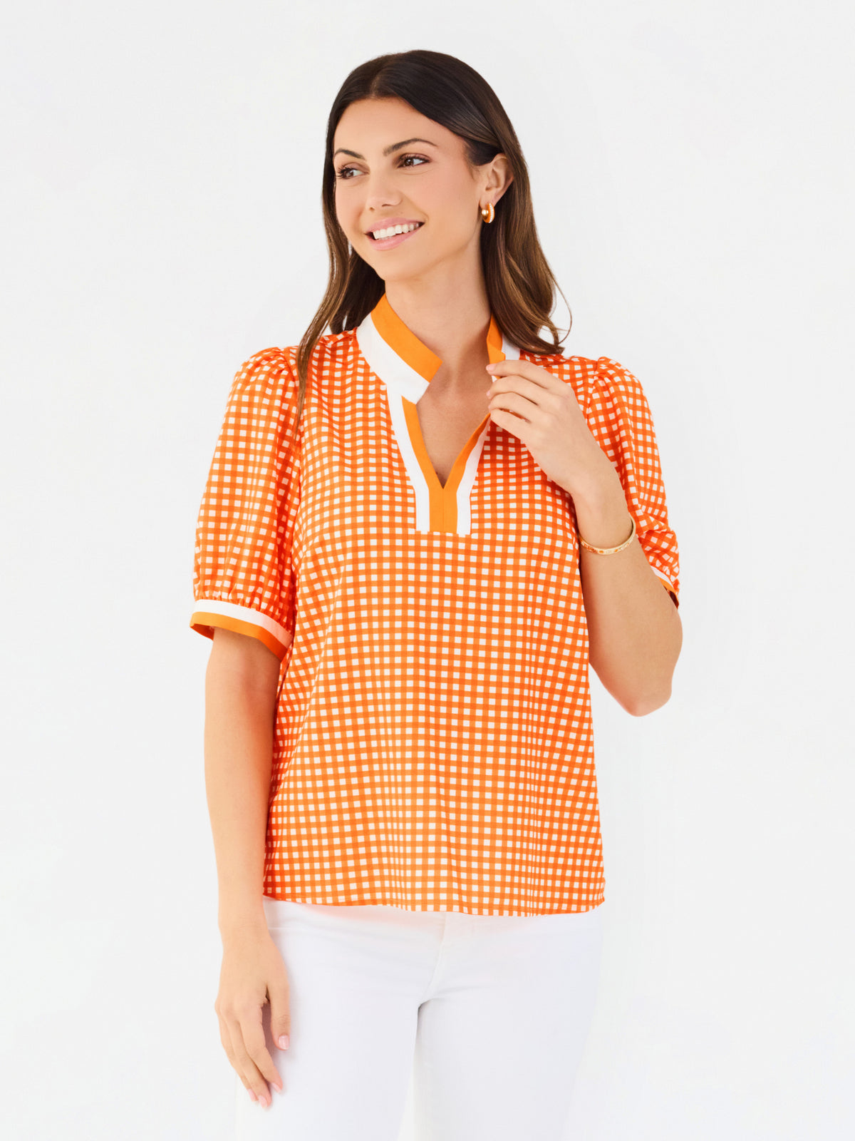 A woman modeling a short-sleeve orange and white gingham top with a white collar and orange trim, paired with white pants, against a plain white background.