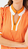 A woman modeling a short-sleeve orange and white gingham top with a white collar and orange trim, paired with white pants, against a plain white background