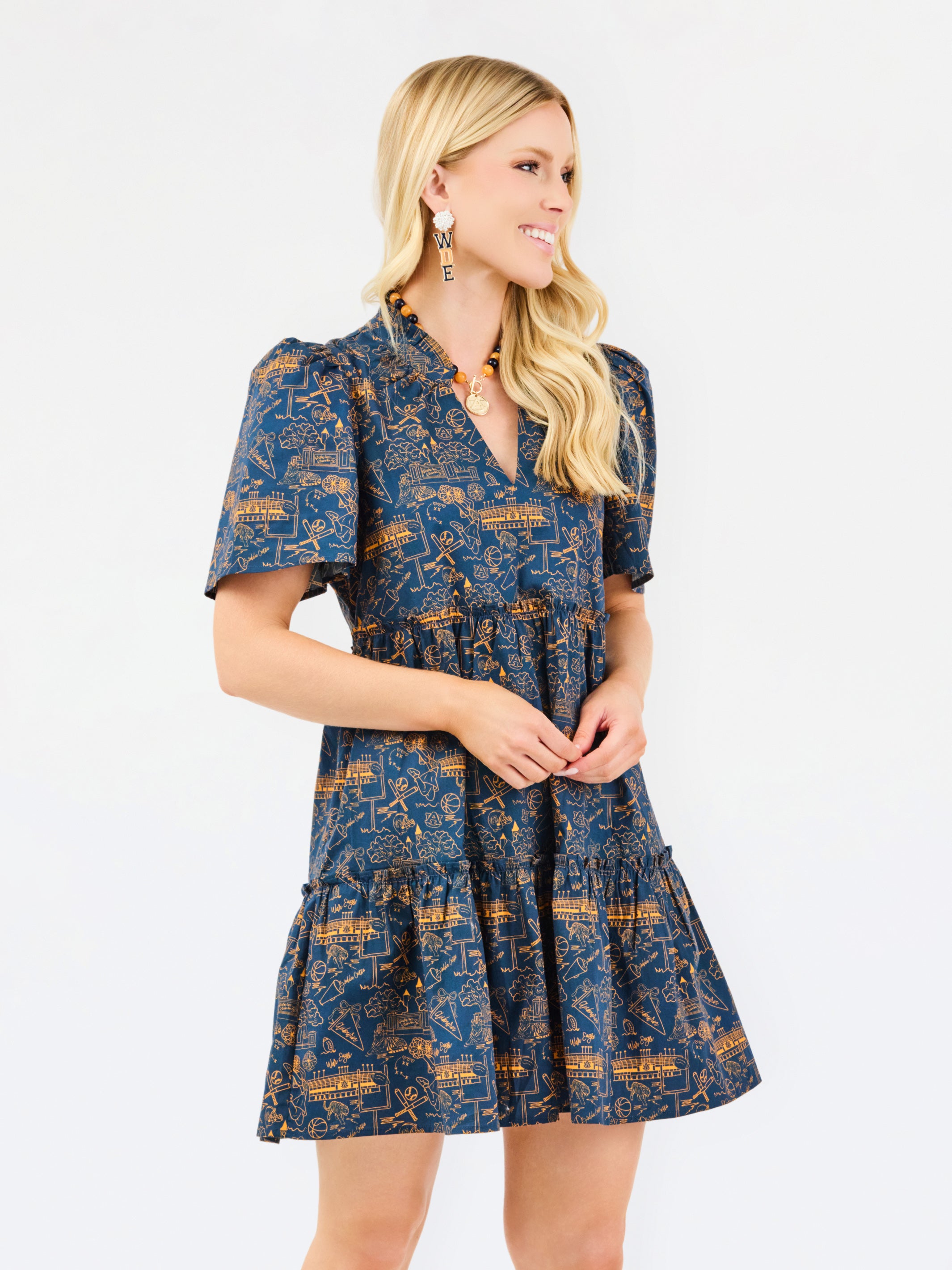 A blonde woman wearing a short-sleeve navy blue dress with a mustard yellow and gold toile pattern, standing against a plain white studio background.