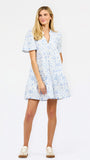 A woman modeling a short-sleeve V-neck dress with a blue and white floral toile pattern, paired with beige sneakers, against a plain white studio background.