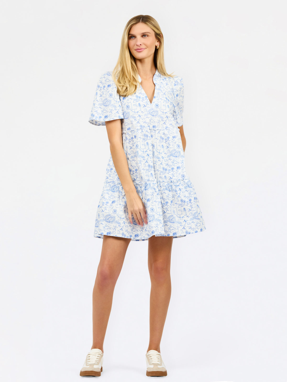 A woman modeling a short-sleeve V-neck dress with a blue and white floral toile pattern, paired with beige sneakers, against a plain white studio background.