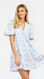 A woman modeling a short-sleeve white dress with a light blue football toile print, featuring a V-neckline and a flared skirt, set against a plain white studio background.