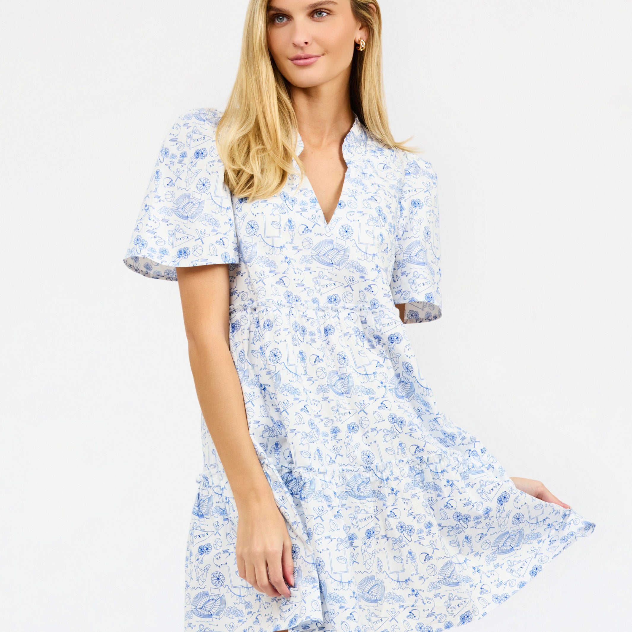 A woman modeling a short-sleeve white dress with a light blue football toile print, featuring a V-neckline and a flared skirt, set against a plain white studio background.
