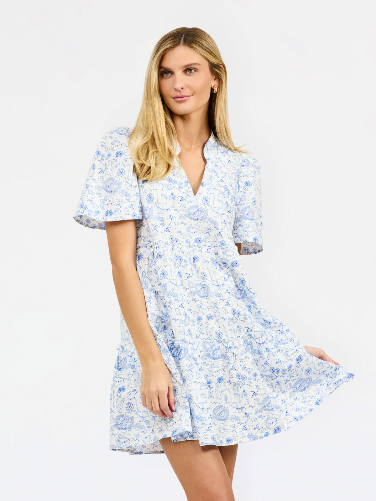 A woman modeling a short-sleeve white dress with a light blue football toile print, featuring a V-neckline and a flared skirt, set against a plain white studio background.