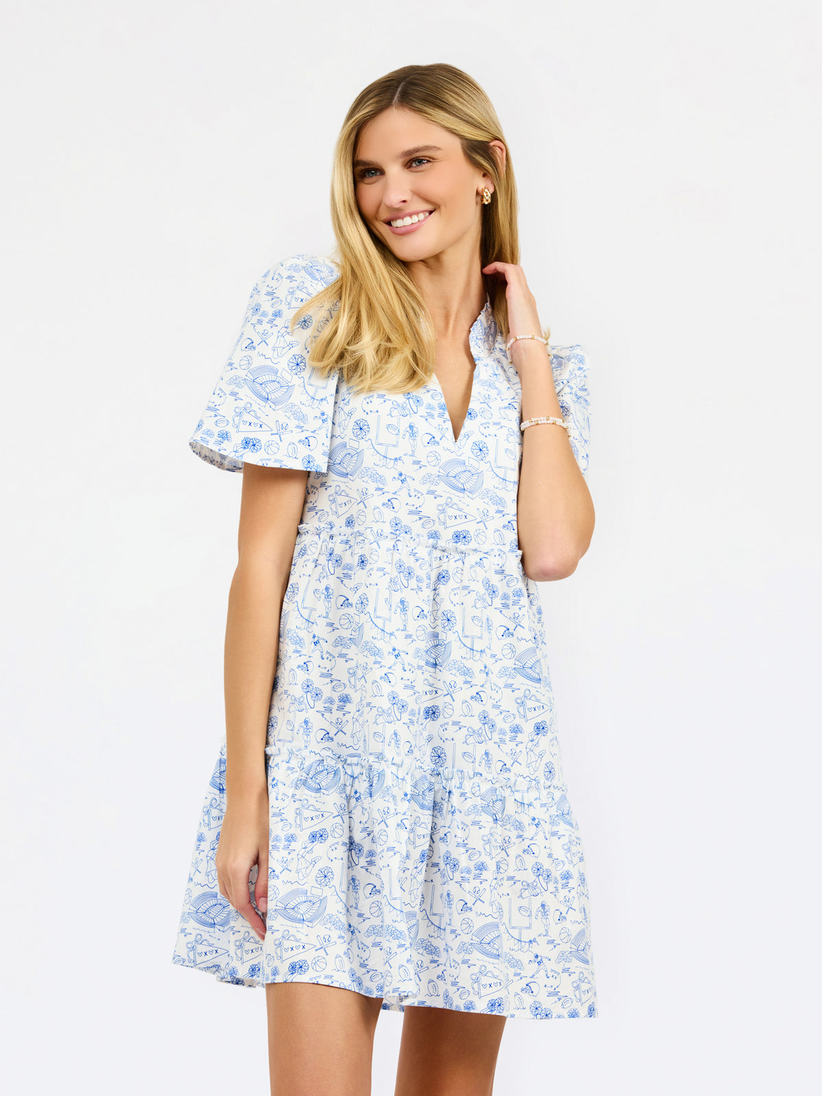 A woman modeling a short-sleeve white dress with a light blue football toile print, featuring a V-neckline and a flared skirt, set against a plain white studio background.