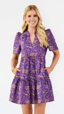 A woman modeling a purple short-sleeve tiered dress with a gold graphic print, standing against a plain white background.
