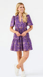 A woman modeling a short-sleeve purple and gold patterned dress with a tiered skirt, paired with white sneakers, against a plain white studio background.