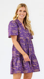 A woman modeling a purple short-sleeve tiered dress with a gold toile-style print, set against a plain white studio background.