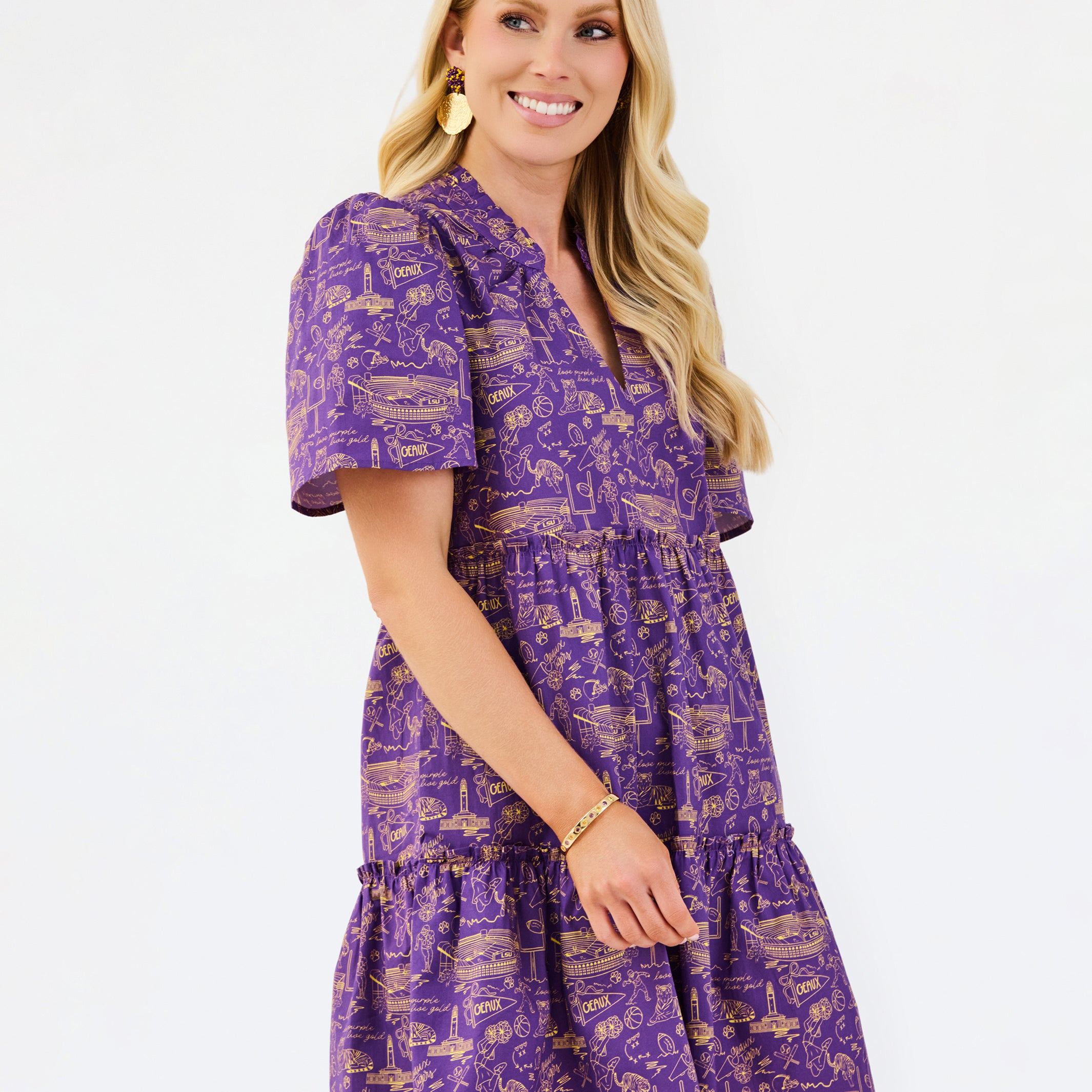 A woman modeling a purple short-sleeve tiered dress with a gold toile-style print, set against a plain white studio background.