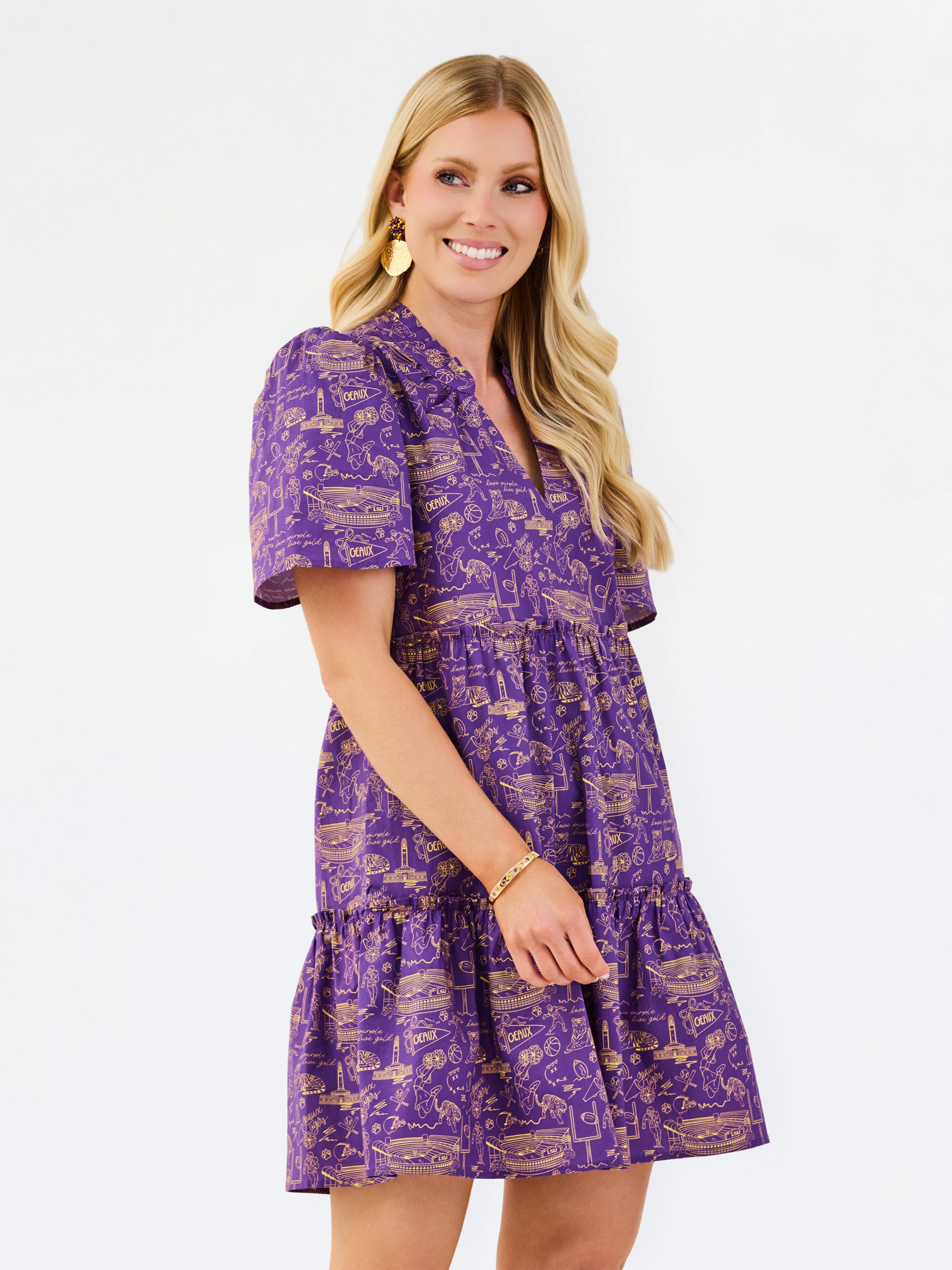 A woman modeling a purple short-sleeve tiered dress with a gold toile-style print, set against a plain white studio background.