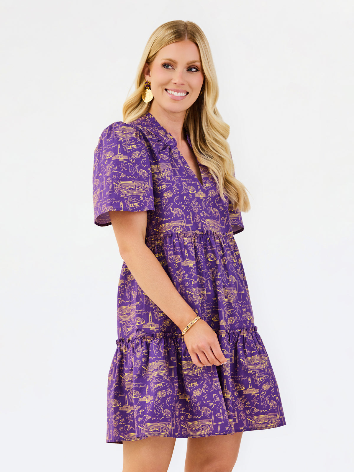 A woman modeling a purple short-sleeve tiered dress with a gold toile-style print, set against a plain white studio background.