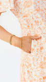 A close-up of a person wearing a white dress with a light orange abstract line pattern and gold beaded bracelets, holding the side pocket of the dress.