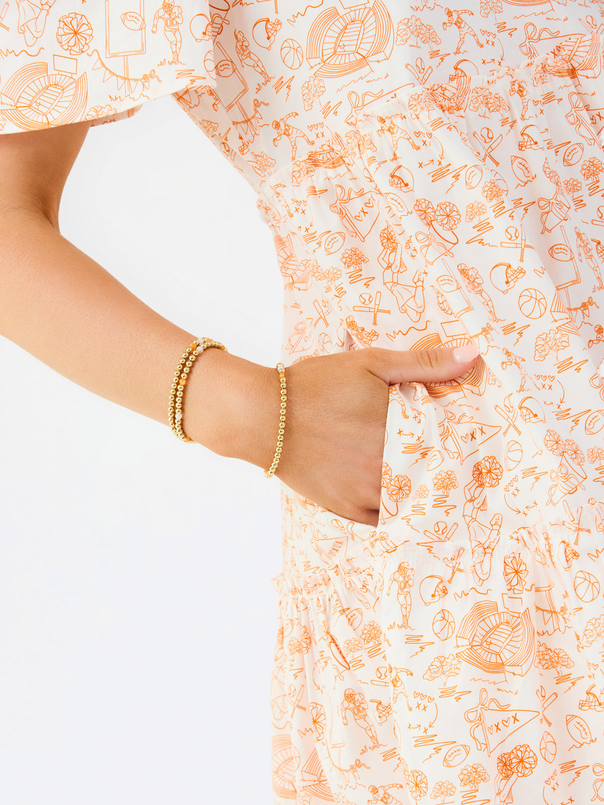 A close-up of a person wearing a white dress with a light orange abstract line pattern and gold beaded bracelets, holding the side pocket of the dress.