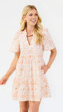 A woman modeling a short-sleeve, tiered mini dress with a peach and orange floral pattern on a white background, set against a plain white studio backdrop.