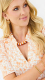 A blonde woman wearing a white short-sleeve blouse with an orange botanical pattern, accessorized with an orange and white beaded necklace and gold bracelets against a plain white background.