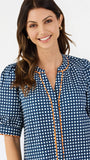 A woman modeling a navy blue and white gingham patterned blouse with a orange trim down the front placket, paired with white pants, against a plain white studio background.