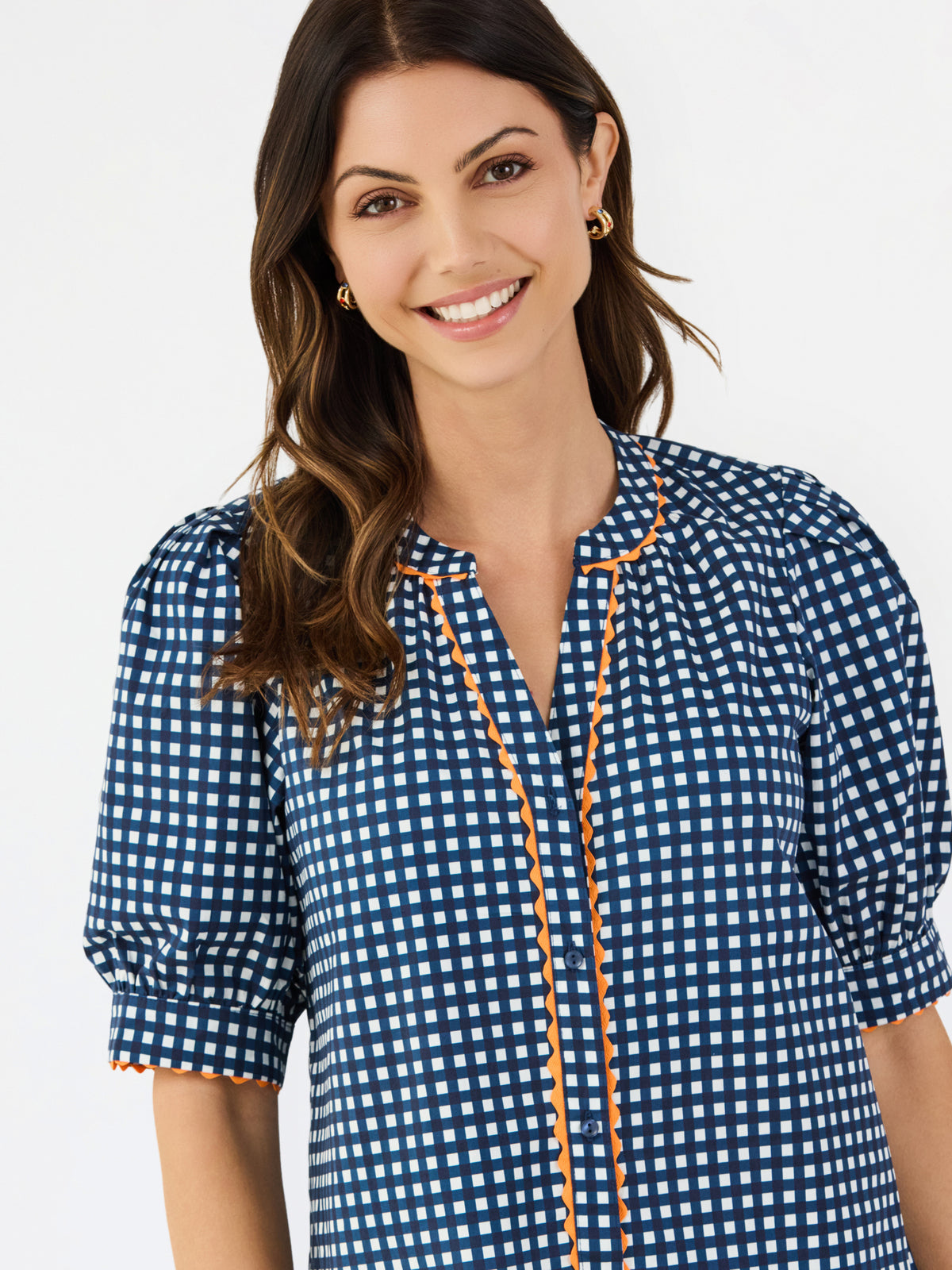 A woman modeling a navy blue and white gingham patterned blouse with a orange trim down the front placket, paired with white pants, against a plain white studio background.