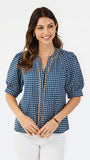 A woman modeling a navy blue and white gingham patterned blouse with a orange trim down the front placket, paired with white pants, against a plain white studio background.