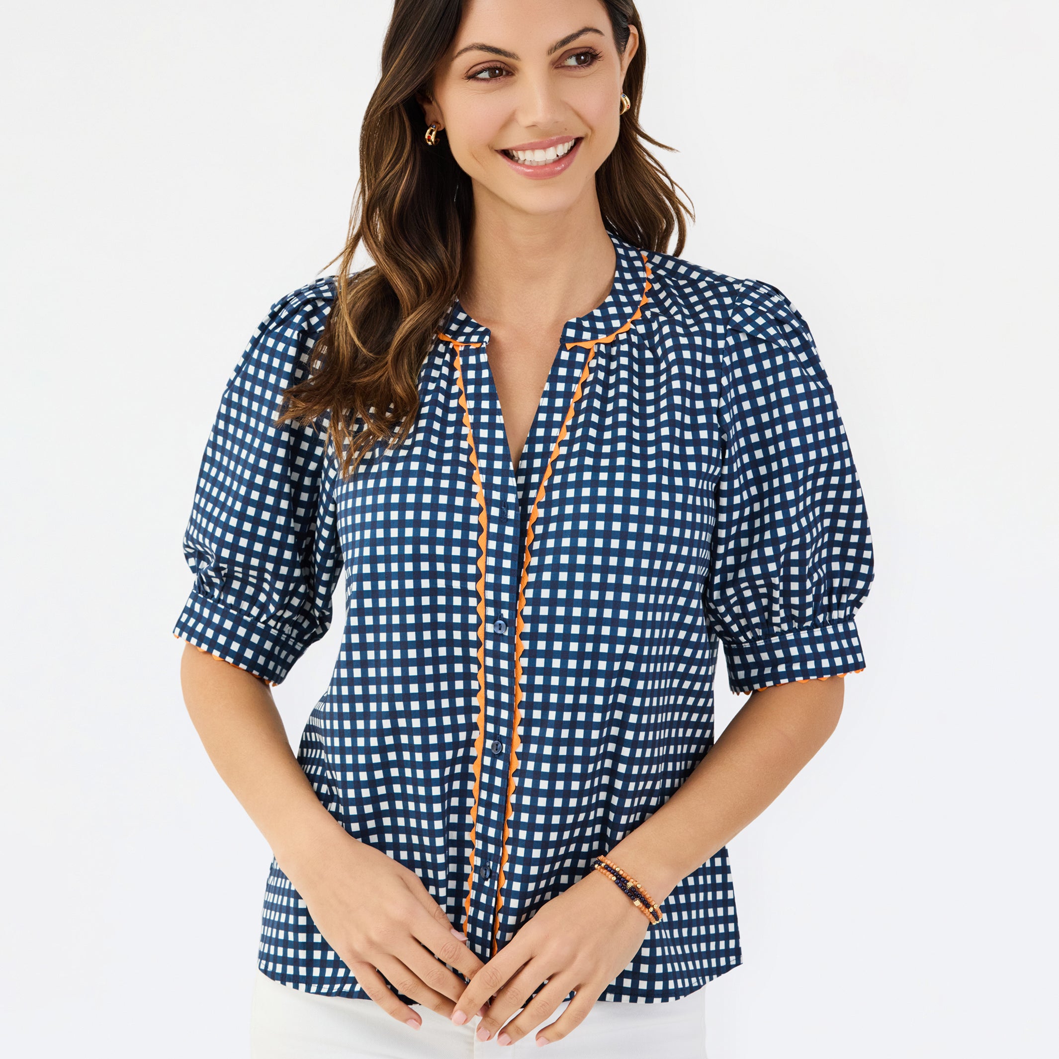 A woman modeling a navy blue and white gingham patterned blouse with a orange trim down the front placket, paired with white pants, against a plain white studio background.