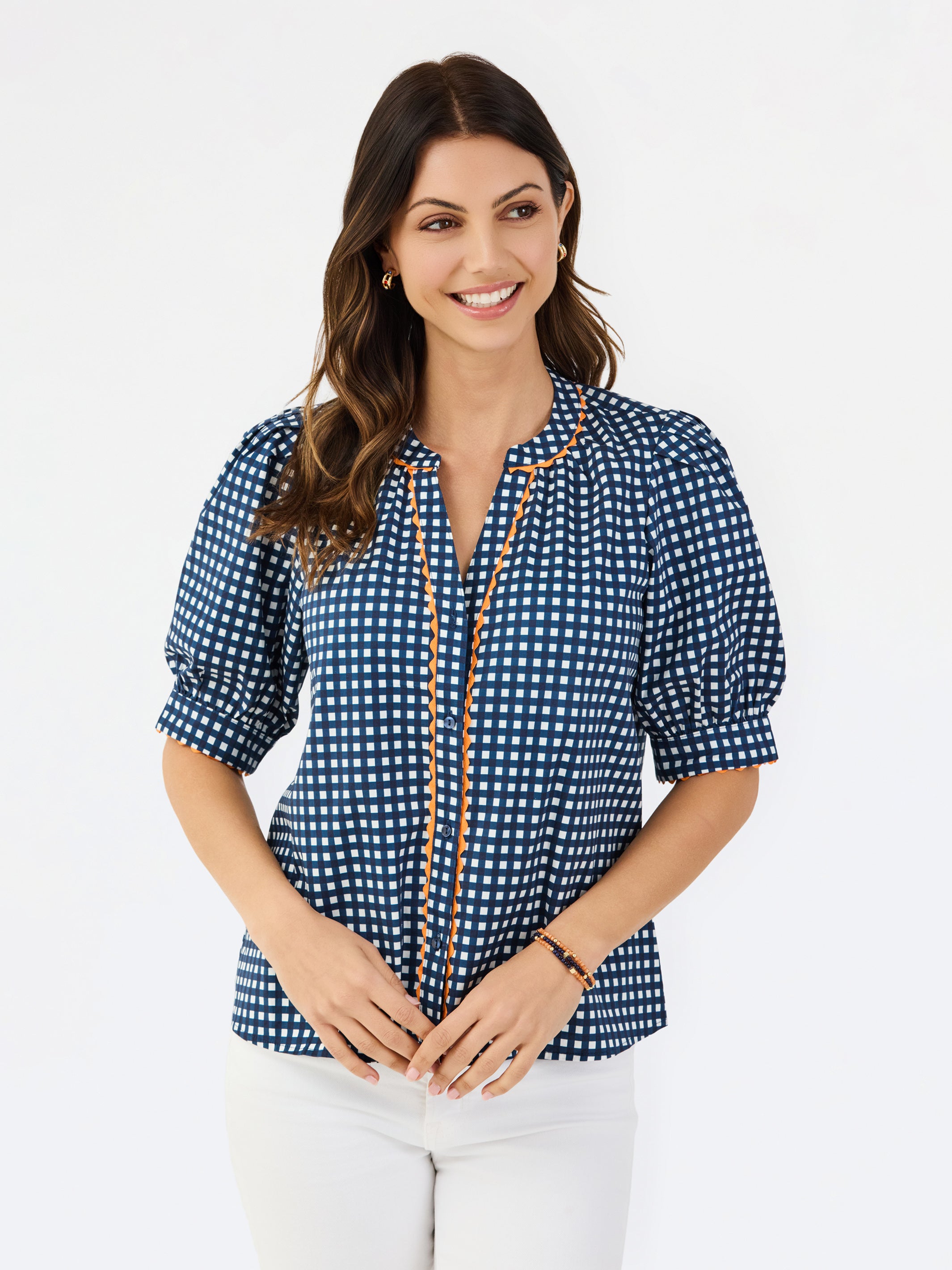 A woman modeling a navy blue and white gingham patterned blouse with a orange trim down the front placket, paired with white pants, against a plain white studio background.