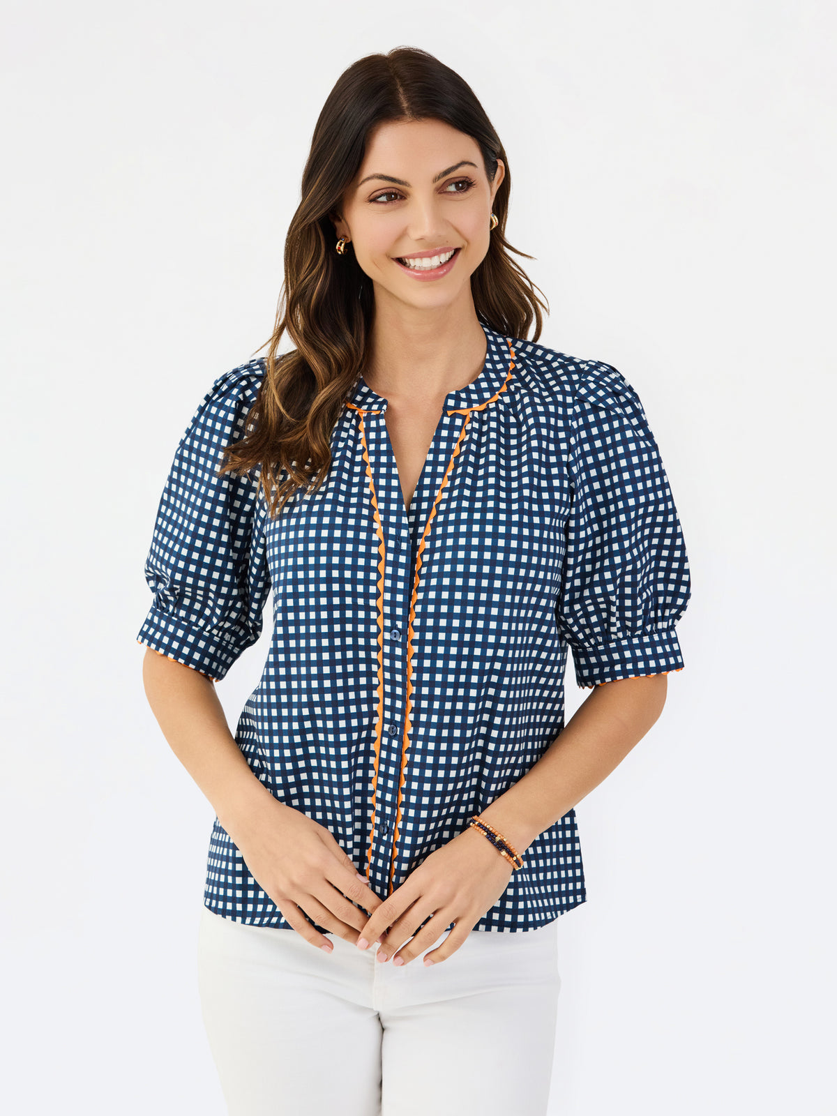 A woman modeling a navy blue and white gingham patterned blouse with a orange trim down the front placket, paired with white pants, against a plain white studio background.