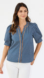 A woman modeling a navy blue and white gingham patterned blouse with a orange trim down the front placket, paired with white pants, against a plain white studio background.