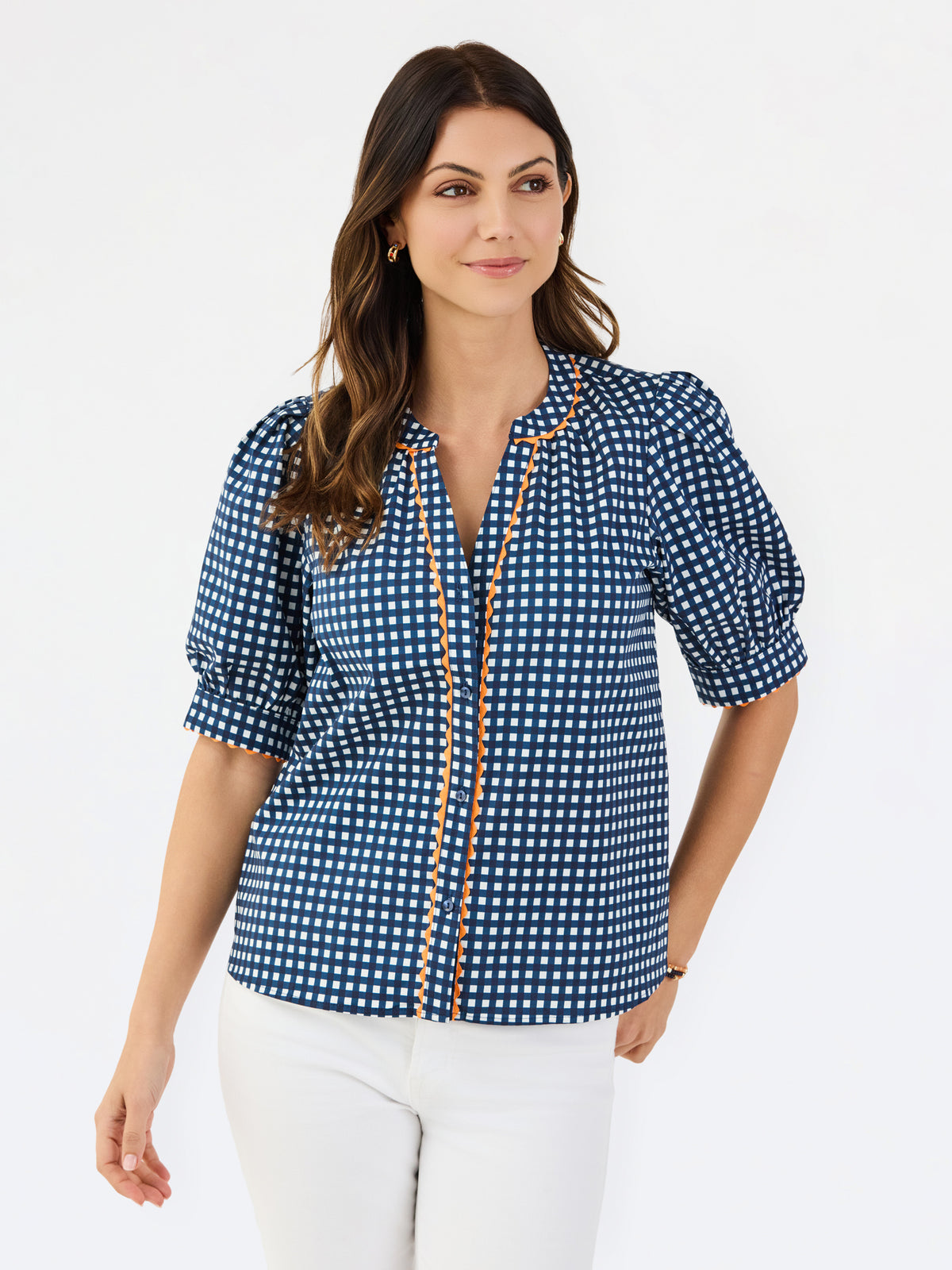 A woman modeling a navy blue and white gingham patterned blouse with a orange trim down the front placket, paired with white pants, against a plain white studio background.