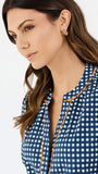 A woman modeling a navy blue and white gingham patterned blouse with a orange trim down the front placket, paired with white pants, against a plain white studio background.