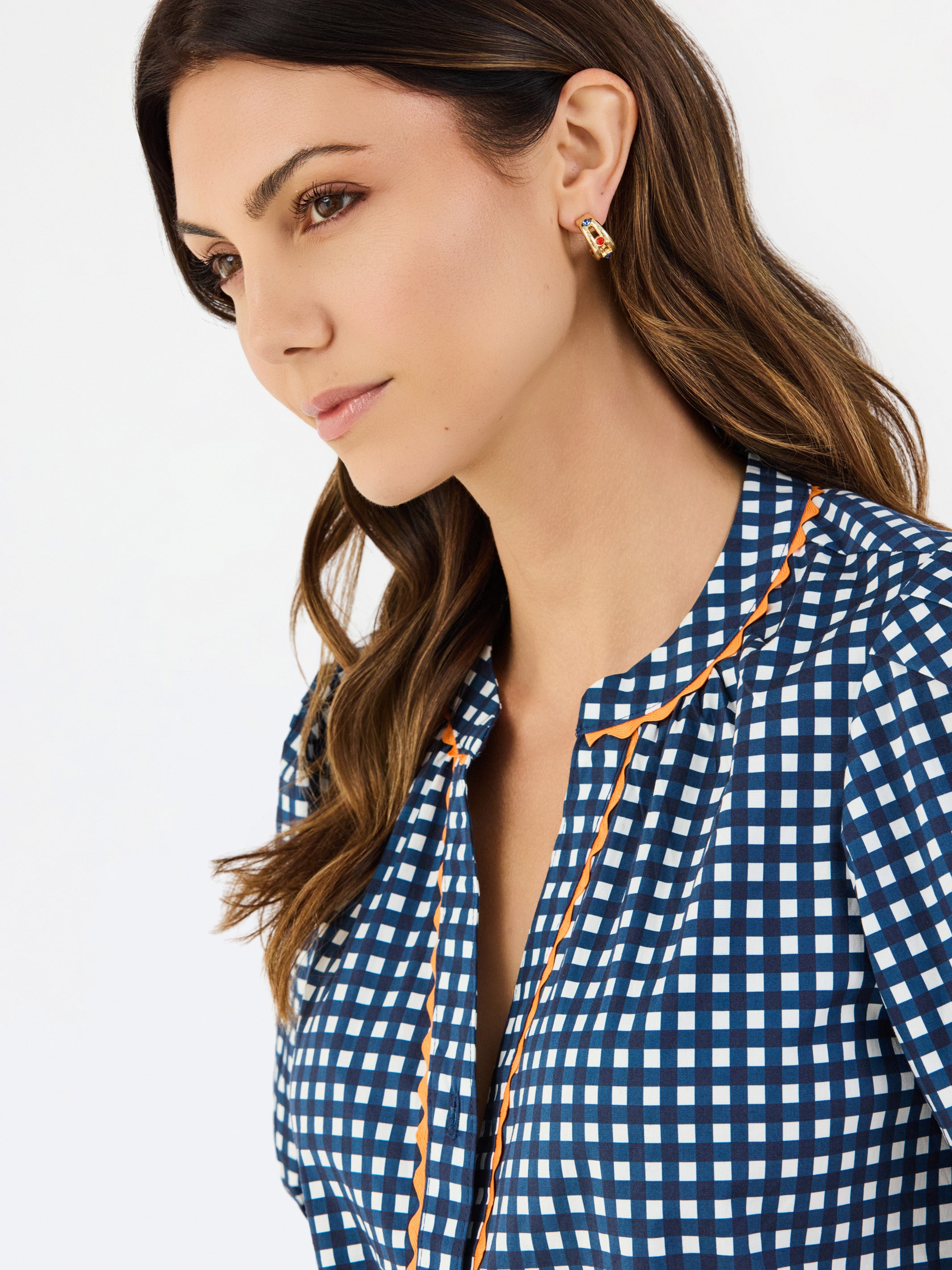 A woman modeling a navy blue and white gingham patterned blouse with a orange trim down the front placket, paired with white pants, against a plain white studio background.
