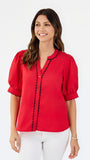 A woman modeling a bright red short-sleeve blouse with black scalloped trim along the button placket and sleeve edges, paired with white pants, against a plain white studio background.
