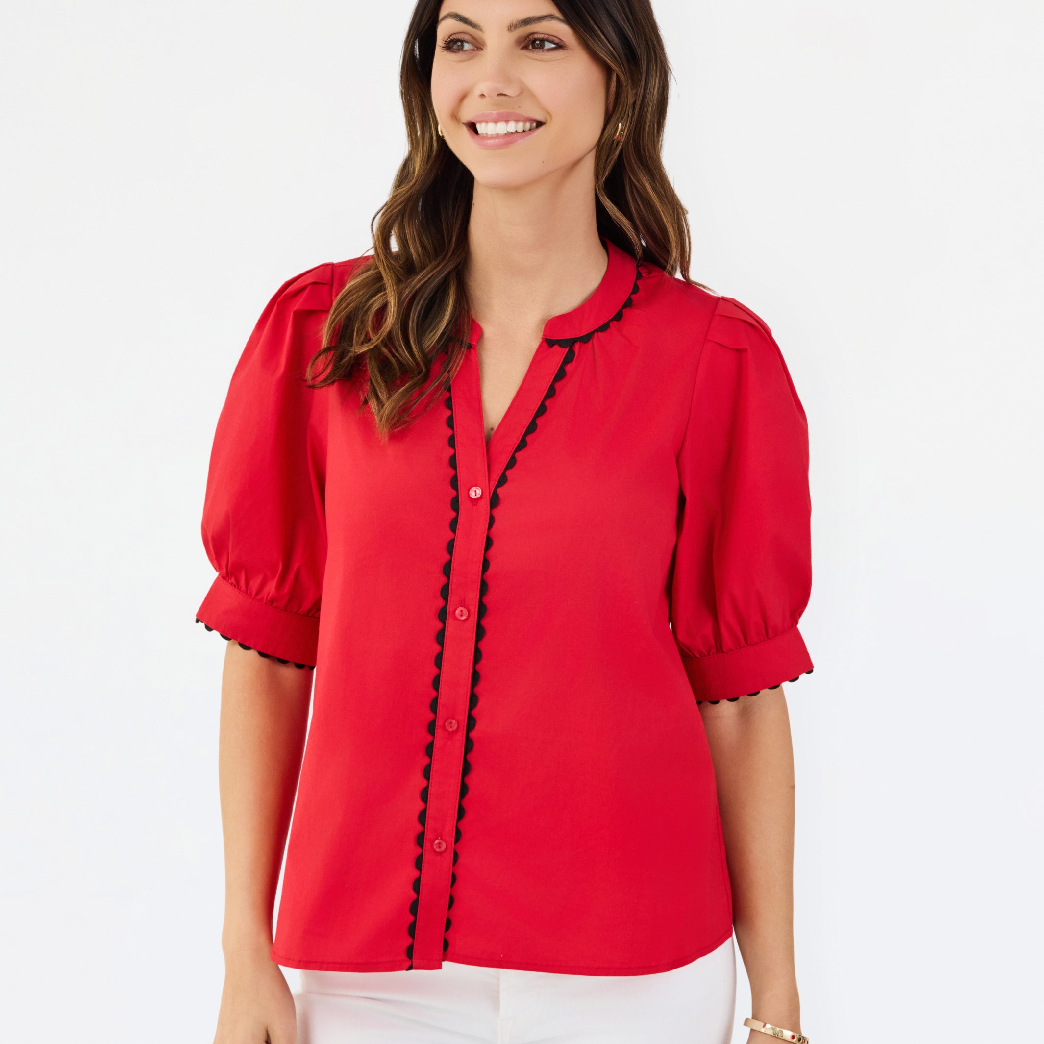 A woman modeling a bright red short-sleeve blouse with black scalloped trim along the button placket and sleeve edges, paired with white pants, against a plain white studio background.