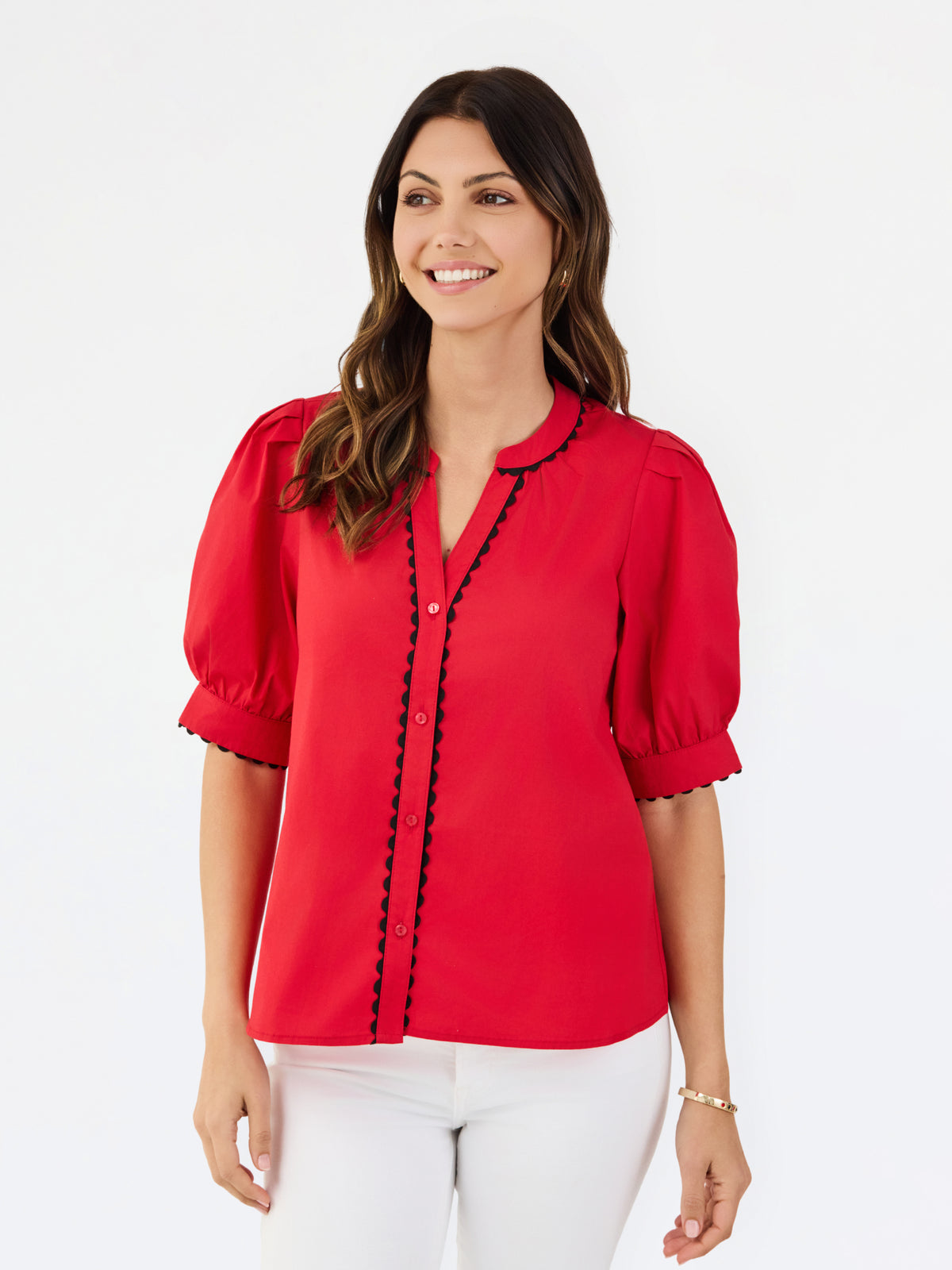 A woman modeling a bright red short-sleeve blouse with black scalloped trim along the button placket and sleeve edges, paired with white pants, against a plain white studio background.