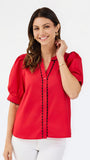 A woman modeling a bright red short-sleeve blouse with black scalloped trim along the button placket and sleeve edges, paired with white pants, against a plain white studio background.