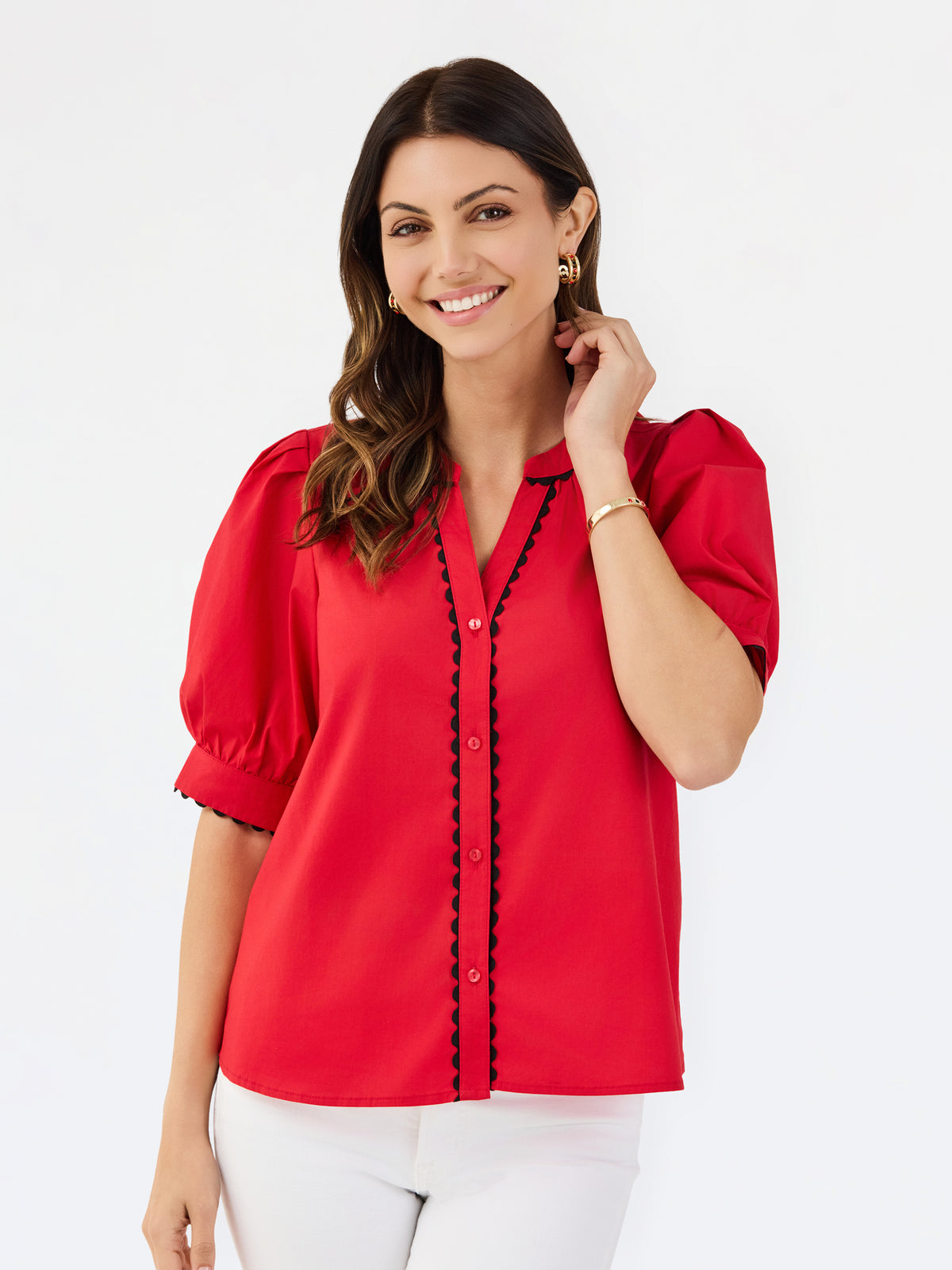 A woman modeling a bright red short-sleeve blouse with black scalloped trim along the button placket and sleeve edges, paired with white pants, against a plain white studio background.